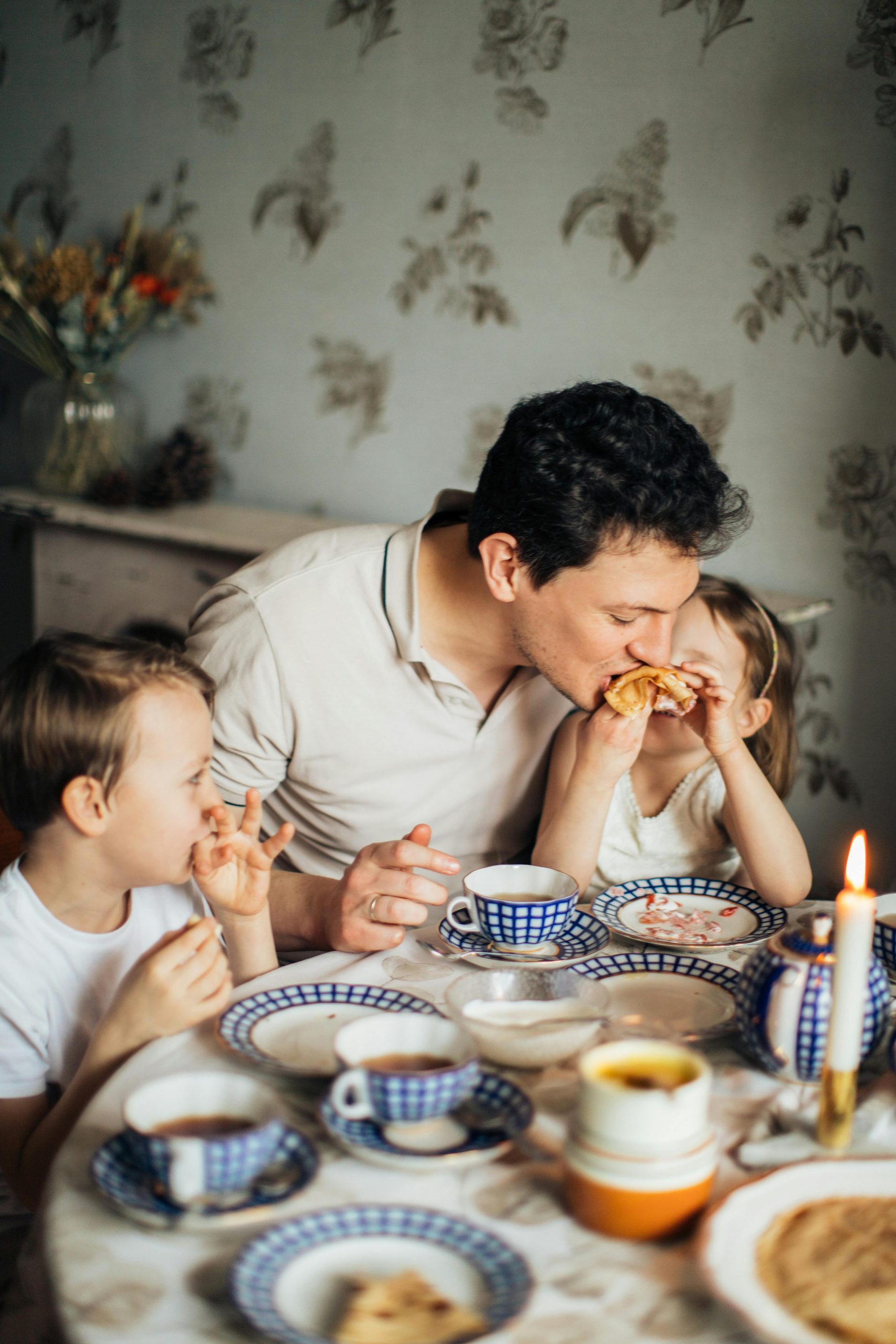 A father and his two children share a joyful meal featuring traditional crepes, creating a warm family moment indoors.
